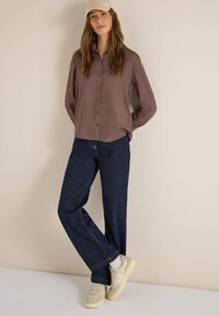 Femme portant une casquette beige, un chemisier marron boutonné, un jean bleu foncé et des baskets beiges, debout contre un mur beige avec les mains dans le dos.