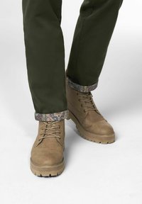 Braune Lederstiefel mit runden Zehen und dicken Sohlen, mit Schnürdesign und einem dekorativen Musterkragen um den Knöchel. Grüne Hose sichtbar.