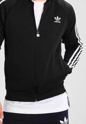 Homme portant une veste zippée noire Adidas avec des bandes blanches sur les manches et le logo Adidas sur la poitrine, par-dessus une chemise blanche.