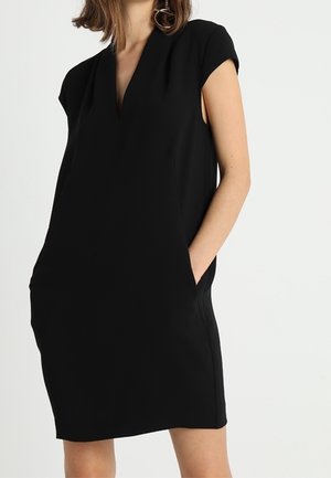 Femme portant une robe noire sans manches, longueur aux genoux, avec un décolleté en V et les mains dans les poches latérales, sur un fond uni.