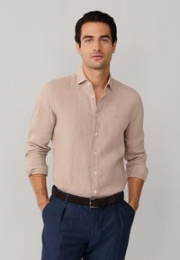 Muškarac obučen u bež košulju dugih rukava s otkopčanim gumbima i uvrnutim rukavima, tamni remen i mornarskoplave plisirane hlače, na jednobojnoj pozadini.