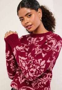 Pulover burgundy cu model floral roz, cu mansete cu nervuri și guler rotund. Fabricat dintr-un material tricotat moale și texturat.