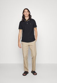 Camisa polo preta com colarinho riscado em amarelo e branco, combinada com calças bege e sandálias pretas. Design simples, textura macia e ajuste casual.