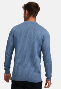 Blå strikket genser med fiskebensmønster, ribbestrikkede mansjetter og hem, rund halsutskjæring og løs passform. Stoffet ser mykt og strukturert ut.