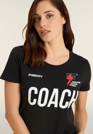 Mujer con cabello castaño hasta los hombros, vistiendo una camiseta negra con los logotipos de "COACH" y "Federazione Italiana Fitness".
