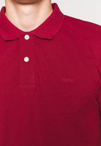 Polo rouge en tissu texturé. Doté d'un col classique, d'une patte de boutonnage à trois boutons et d'un logo en relief discret sur la poitrine.