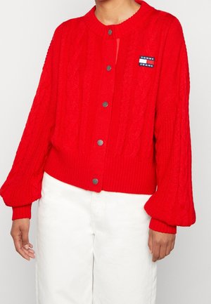 Rode kabelgebreide cardigan met zilveren knopen en Tommy Jeans logo, gedragen met witte broek, getoond van nek tot heupen.