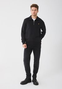 Felpa nera con zip e colletto, dotata di un piccolo logo giallo, abbinata a jeans scuri e stivali neri con lacci su uno sfondo neutro.