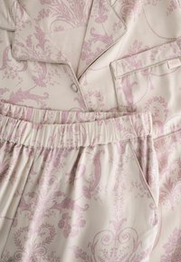 Pyjamapukuna, joka sisältää vaaleanbeigeä kangasta, jossa on monimutkaisia vaaleanpunaisia kukkakuoseja, kauluksellisen paidan ja elastiset vyötäröhousut.