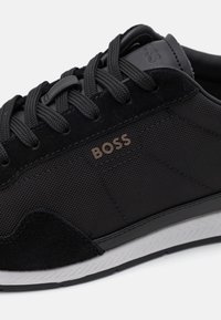 Svart sneakers laget av teksturert stoff og semsket skinn, med svarte lisser, gummisåle og en subtil "BOSS"-logo preget på siden.