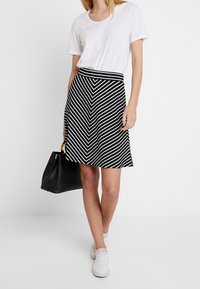 Hame haji mustri hitam dan putih dengan desain mengembang, panjang di atas lutut, dipadukan dengan kaos putih polos dan sepatu sneaker putih. Termasuk tas tangan hitam.