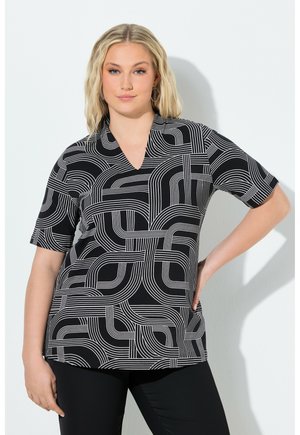 Blond kvinde iført en sort-hvid top med geometrisk mønster og korte ærmer med V-udskæring, poserende med hånden på hoften foran en hvid væg.