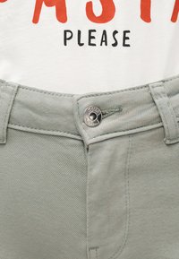 Κοντινό πλάνο ελαφρώς πράσινων τζιν με κουμπωμένο το κουμπί στη μέση και λευκό πουκάμισο μέσα, που δείχνει τη λέξη «PLEASE» με μαύρα και κόκκινα γράμματα.