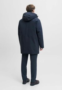 Tamsiai mėlynas vandeniui atsparus striukė su gobtuvu, tekstūruota medžiaga, tiesus kirpimas ir užslėpta užtrauktukas. Dėvima virš pilkų kelnių ir tamsių batų.