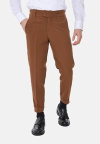 Pantaloni tailored marroni con una texture liscia, dotati di chiusura con bottone laterale, gambe affusolate e pieghe frontali, abbinati a scarpe nere.