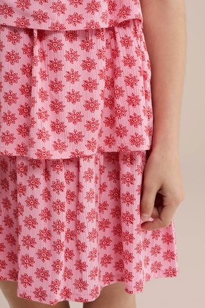 Primo piano di un vestito rosa con motivi floreali rossi e gonna a balze, con un braccio e una mano parzialmente visibili.