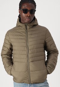 Olivengrüne, gepolsterte Jacke mit horizontalen Nähten, vorderem Reißverschluss und passender Kapuze. Getragen mit blauen Jeans und dunklen Sonnenbrillen.