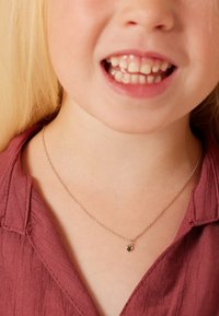 Zilveren ketting met een kleine harthanger, gedragen door een kind. Het topje is maroon van kleur en heeft een gemêleerde stof.
