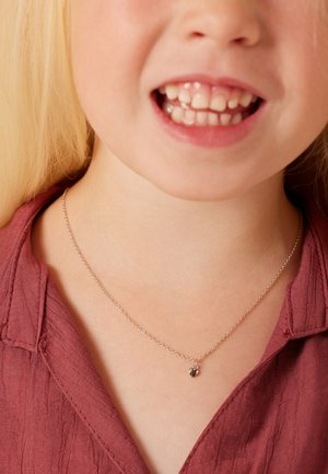Collier en chaîne argentée avec un petit pendentif en forme de cœur, porté par un enfant. Le haut est de couleur bordeaux avec un tissu texturé.