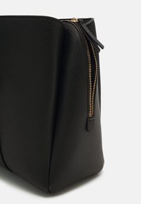 Pochette en cuir noir texturé avec une fermeture éclair dorée partiellement ouverte, montrant une forme rectangulaire avec un bord supérieur légèrement incurvé.