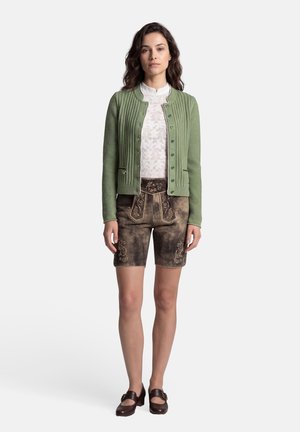 Femme portant un cardigan vert boutonné, une blouse blanche en dentelle, un short en cuir marron brodé, et des chaussures marron foncé à boucle, debout face à l'avant.