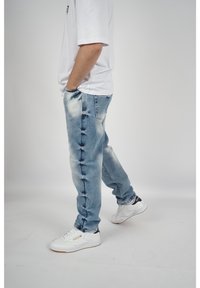 Jeans de mezclilla azul claro con una textura desgastada y deshilachada, combinados con zapatillas blancas. Diseño ajustado con bolsillos visibles en los laterales.