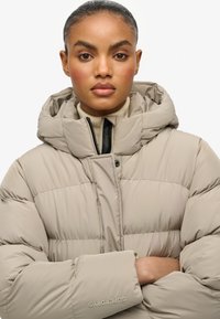 Chaqueta de plumas beige con capucha, con un cierre frontal con cremallera, textura acolchada y logotipo bordado en la manga. Tejido liso y ligero.