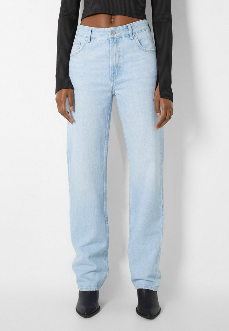 Femme portant un jean droit taille haute bleu clair, des bottes noires pointues et un top court noir à manches longues, debout devant un fond blanc.