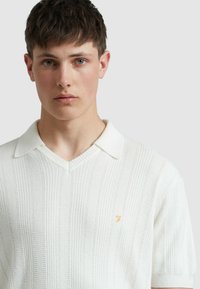 Weißes gestricktes Polohemd mit V-Ausschnitt, gerippter Textur, kurzen Ärmeln und einem kleinen goldenen Logo auf der oberen linken Seite.