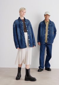 To modeller står mot en nøytral bakgrunn. Kvinnen har på seg en quiltet denimjakke over en brun strikket topp og en hvit ribbestrikket skjørt. Hun har svarte ankelstøvler. Mannen har på seg en lignende denimjakke over en beige skjorte og mørke denimbukser, med svarte sko.