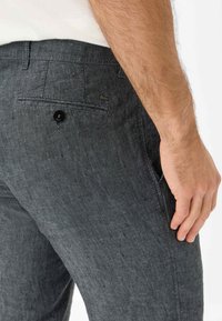 Pantaloni in lino grigio scuro da uomo con tasca posteriore, chiusura con un singolo bottone nero e una leggera texture. Etichetta del logo visibile sulla vita.