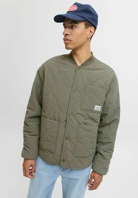Gequiltede olijfgroene jas met drukknoopsluiting en een borstzak, gedragen over lichtblauwe jeans. Vergezeld van een marineblauwe pet met een logo.