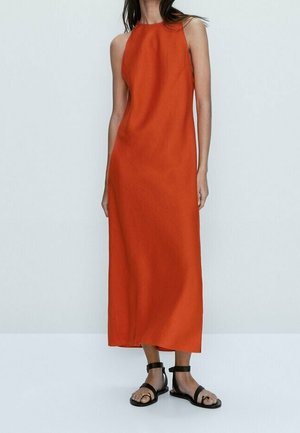 Woman wearing a sleeveless, ankle-length orange dress with black ankle-strap flat sandals, standing against a plain light background.