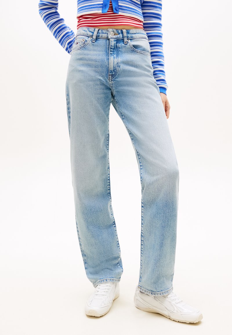 Άτομο που φοράει ανοιχτό μπλε τζιν με ίσια γραμμή, λευκά αθλητικά παπούτσια και κοντό μπλουζάκι με μπλε και κόκκινες ρίγες, στέκεται μπροστά σε λευκό φόντο.