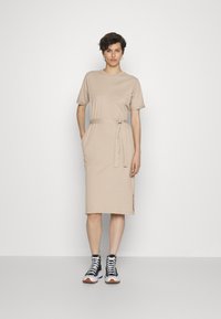 Vestido de algodão bege com mangas curtas, cintura cintada, bolsos laterais e comprimento até ao joelho. Combinado com sapatilhas de cano alto em preto e branco.