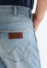 Φωτεινό γαλάζιο τσέπα τζιν Wrangler με καφέ δερμάτινο λογότυπο και ένα χέρι που ακουμπά στην μπροστινή τσέπη πάνω σε απλό φόντο.