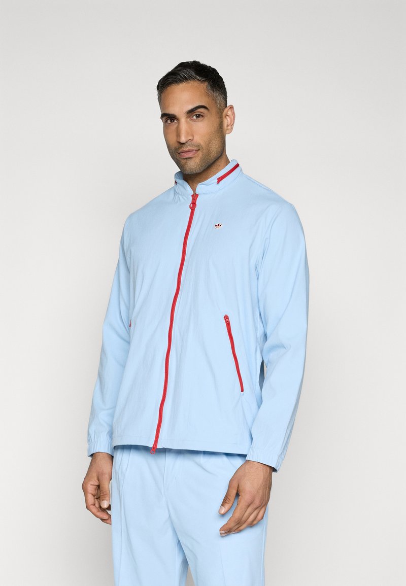 Chaqueta azul claro con cremallera y acentos rojos, cuello tipo levantado y bolsillos laterales. Textura suave con un corte relajado.