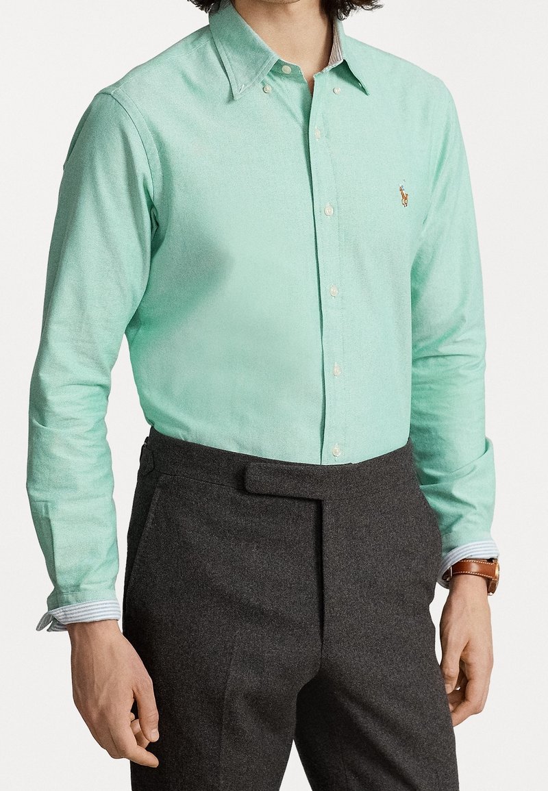 Ljusmintgrön skjorta med knappslå och en subtil textur, krage och broderad logotyp på bröstet; bärs med mörkgrå byxor och randig manschett.