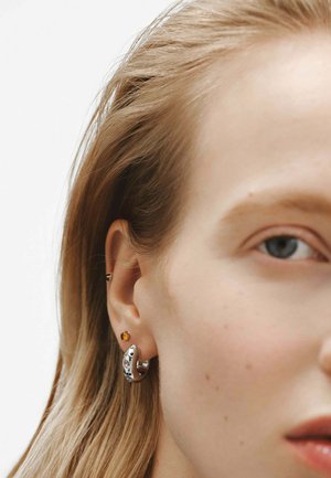 Pendiente de aro plateado con piedras de acento azules, acompañado de un pequeño pendiente dorado en la oreja. El fondo es liso y neutro. El cabello es liso y claro.