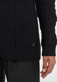 Maglione nero lavorato a maglia con punti a rilievo, polsini a costine e bottone con logo nero di lato. Il materiale appare spesso e accogliente.