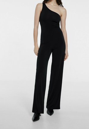 Kvinde iført en sort jumpsuit med én skulder, vide ben og sorte spidse sko mod en ensfarvet hvid baggrund.