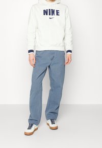 Vit hoodie med marint "NIKE"-logotyp, kombinerad med ljusblå jeans och vita sneakers med marina snören och en beige sula. Enkelt design.