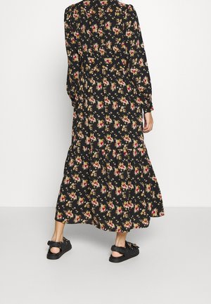 Czarna maxi sukienka z długim rękawem w czerwone, żółte i beżowe kwiatowe wzory, noszona z czarnymi sandałami, pokazana od tyłu na białym tle.