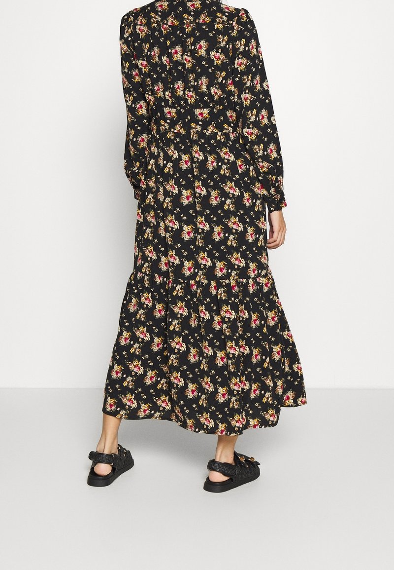 Robe longue noire à manches longues avec imprimé floral rouge, jaune et beige, portée avec des sandales noires, vue de dos sur un fond blanc.