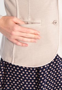 Beżowa marynarka o gładkiej fakturze, zapinana na guziki, z boczną kieszenią, nałożona na ciemną spódnicę w kwiatowy wzór.