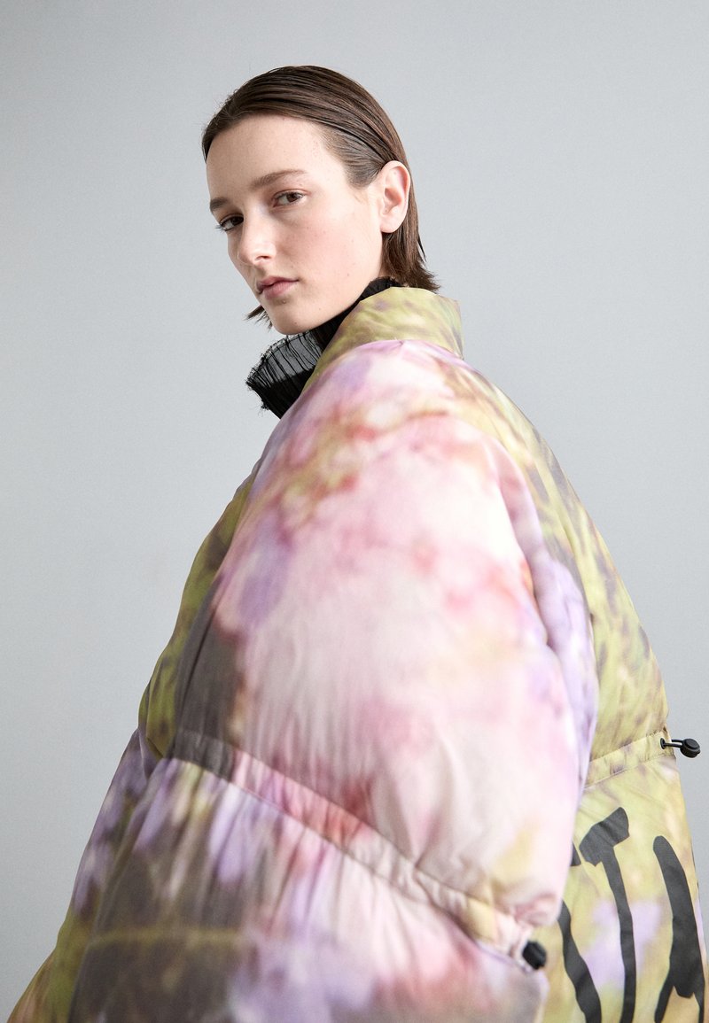 Pružná bunda v pastelových barvách s květinovým vzorem, která má vysoký límec a černé kovové detaily. Texturovaná tkanina s lesklým povrchem.