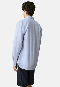 Camicia a maniche lunghe a righe blu chiaro con polsini abbottonati, dettagli con piega sul retro e orlo arrotondato, abbinata a pantaloni corti scuri.