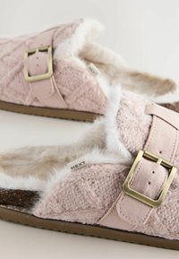 Rosa, flauschige Hausschuhe mit einem strukturierten Obermaterial, seitlicher Schnallenhardware und einem weichen Fellfutter. Die Sohle ist braun und leicht strukturiert.