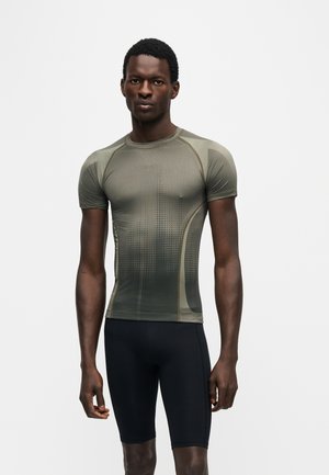 Bărbat stând în fața unui fundal simplu, purtând un tricou sport mulat verde măsliniu și pantaloni scurți negri de ciclism, cu fața în față.