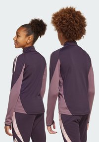 Veste de sport violette avec des accents roses, dotée d'un col haut et de manches longues. Texture lisse avec des panneaux contrastants sur les côtés.